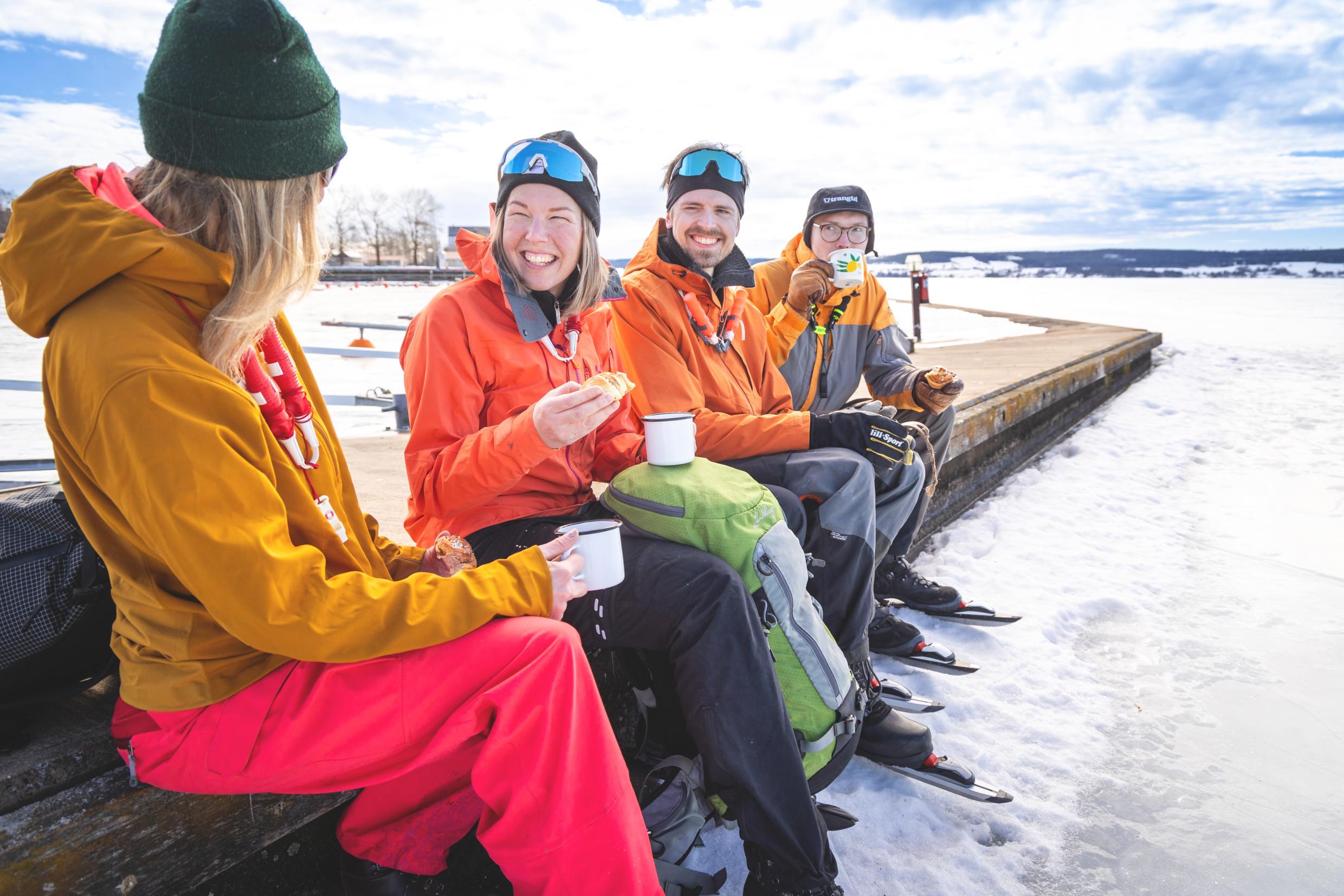 A group of four drinking coffee next to an ice-covered lake. They are wearing ice skates.